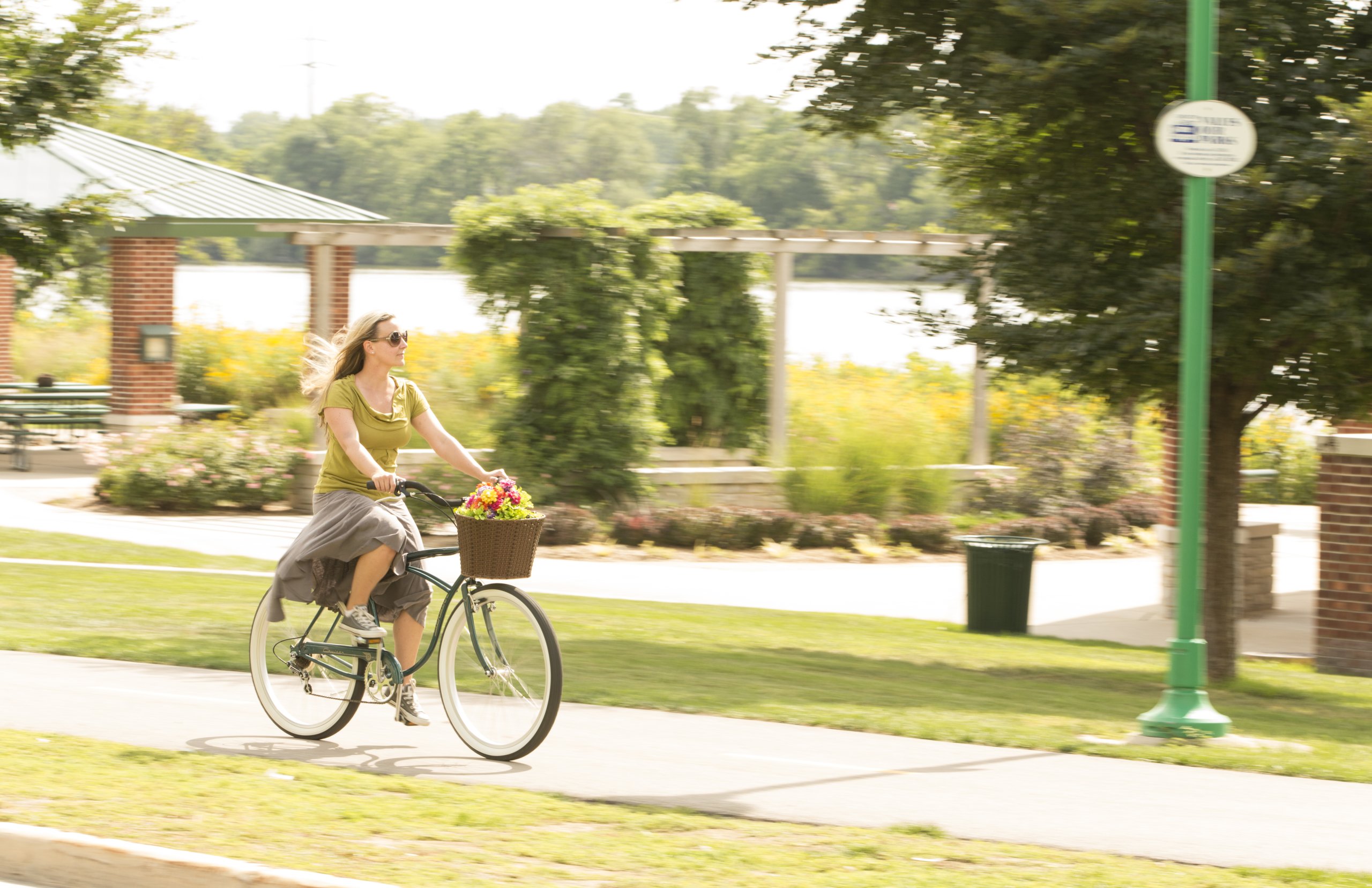 A woman rides a bike with a basket full of flowers along a bike path in a park on a sunny day.