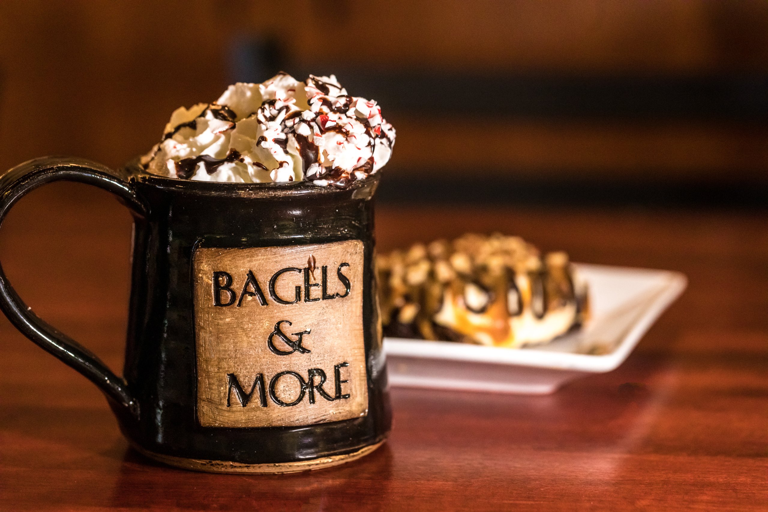 Bagels and More coffee cup is topped with whipped cream with chocolate drizzle and peppermint crumbs sits in front of a plate of pastry.